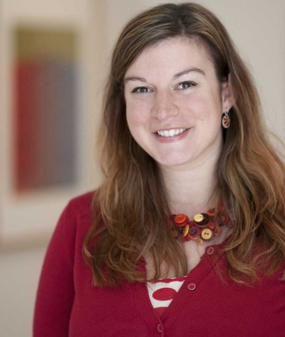 A woman with light brown hair smiles at the camera. She is wearing a red sweater and a necklace made of orange and brown buttons.