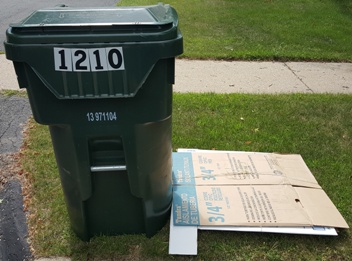 Stack of excess cardboard cut down to 3 foot by 3 foot squares, bundled with twine, and neatly stacked next to the recycling cart